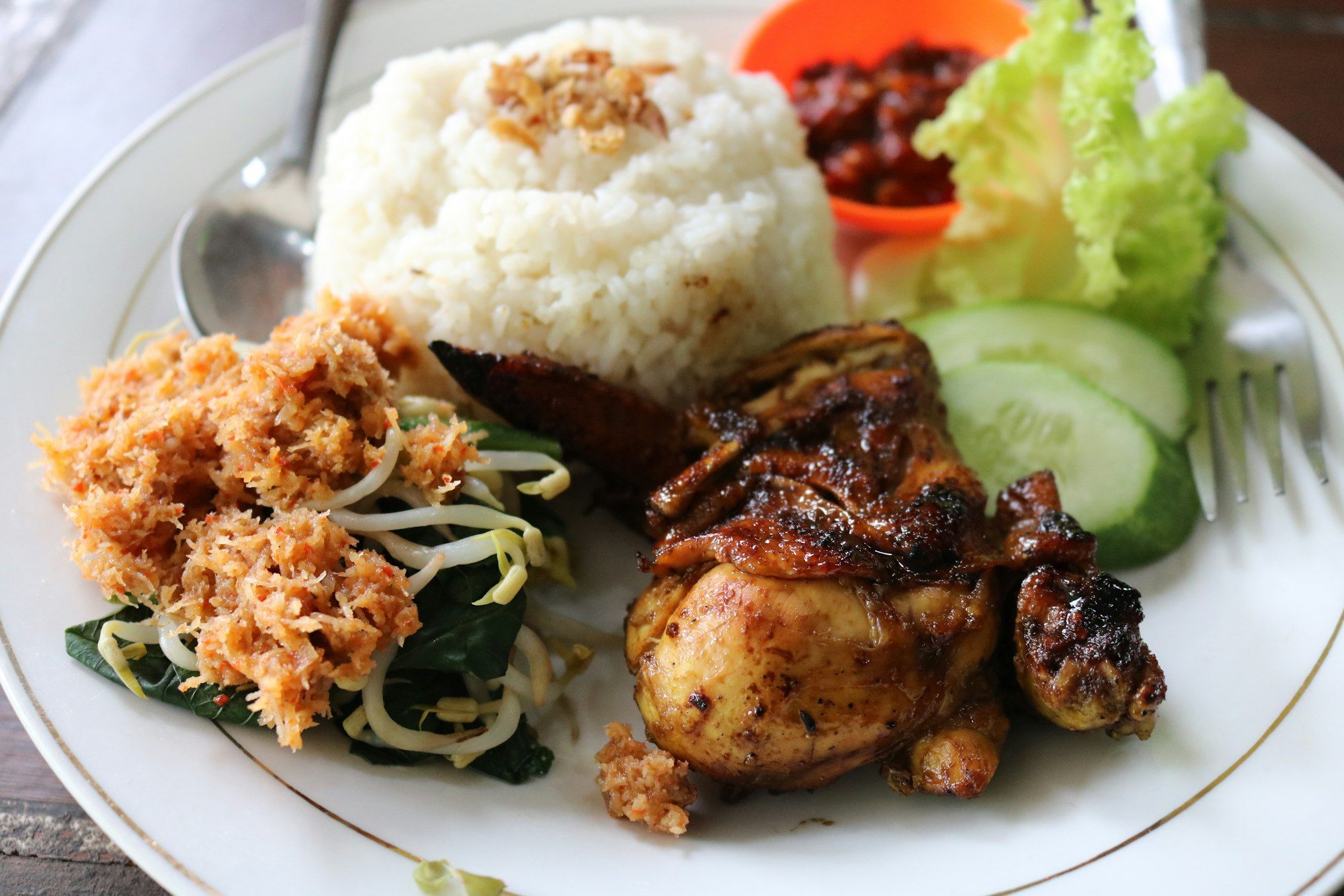 Piring berisi nasi, ayam bakar, urab tauge dan daun, irisan timun, serta sambal di mangkuk oranye