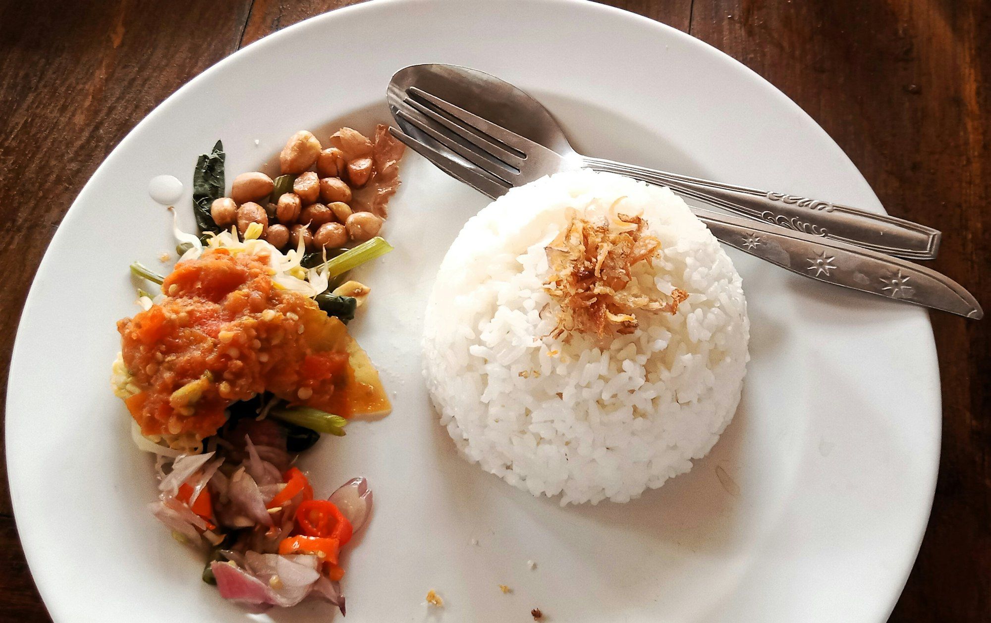 Piring putih berisi nasi hangat dengan bawang goreng, kacang goreng, tahu bersambal tomat, dan sambal matah