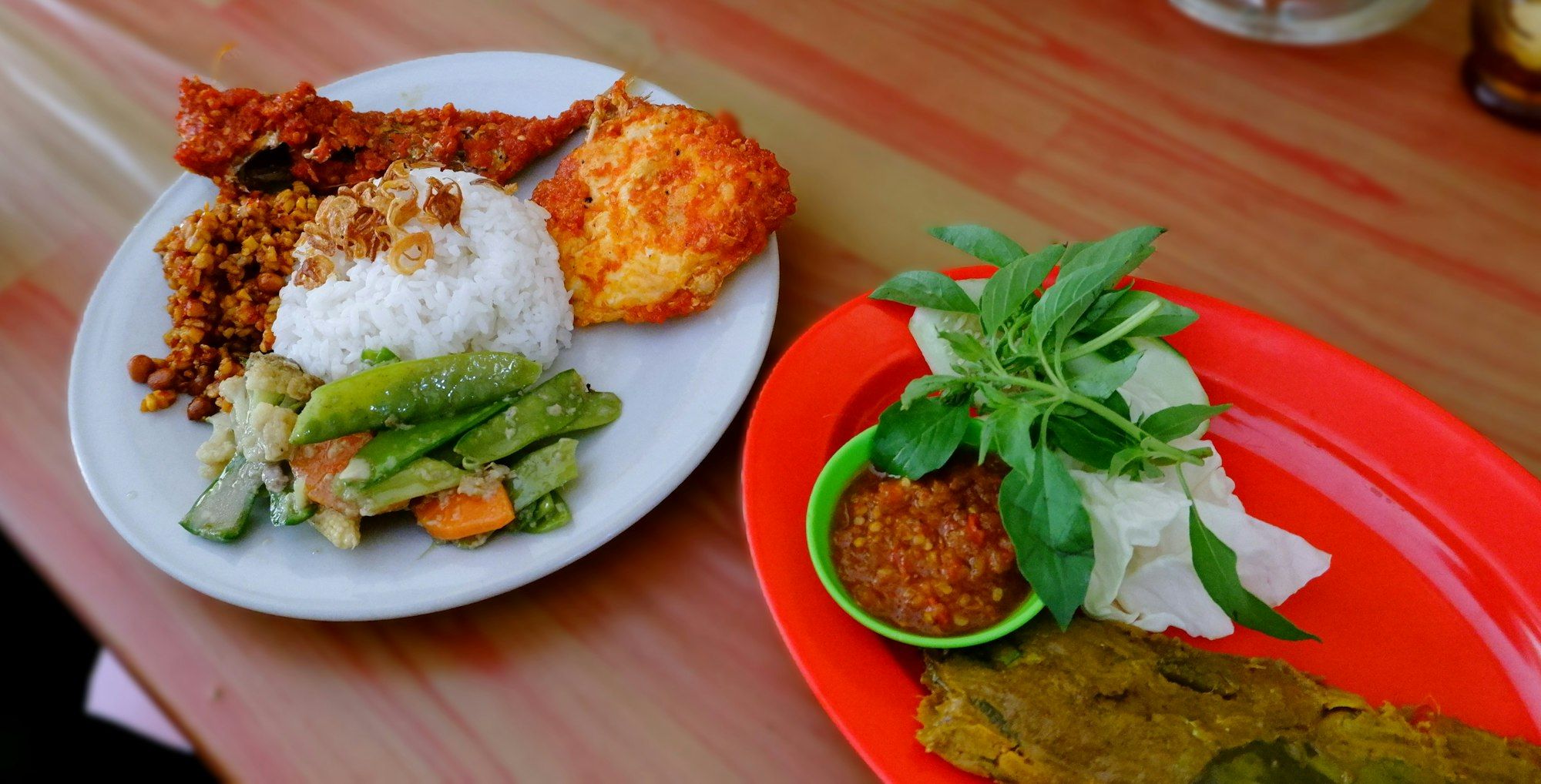 Dua piring: nasi dengan tempe orek, ikan bersambal, telur, dan sayur; di sebelahnya sambal, kemangi, dan kol segar
