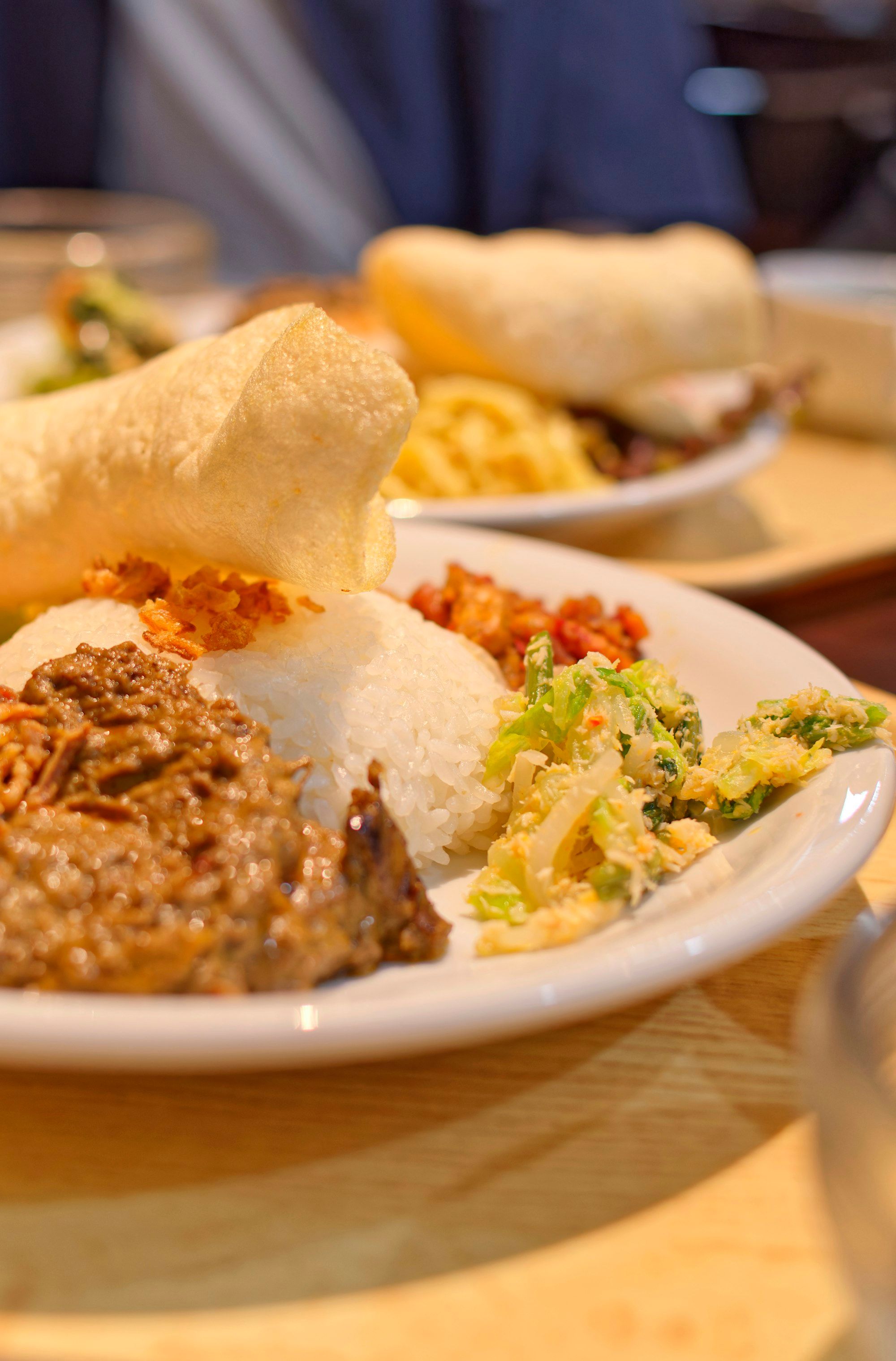 Piring nasi dengan daging berbumbu cokelat pekat, urab sayur, dan kerupuk udang