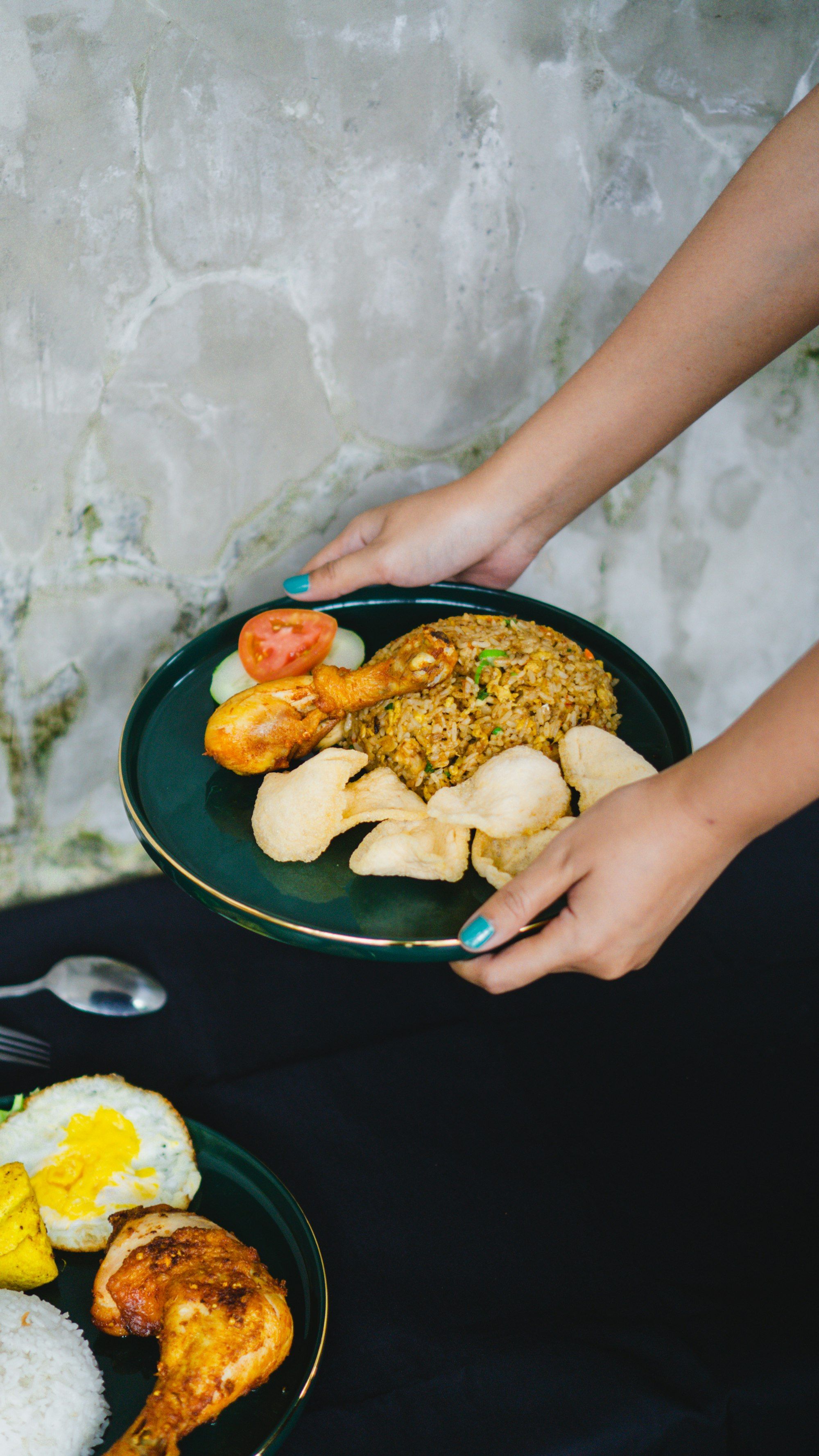 Sepasang tangan menaruh piring nasi goreng berisi ayam dan kerupuk di atas meja