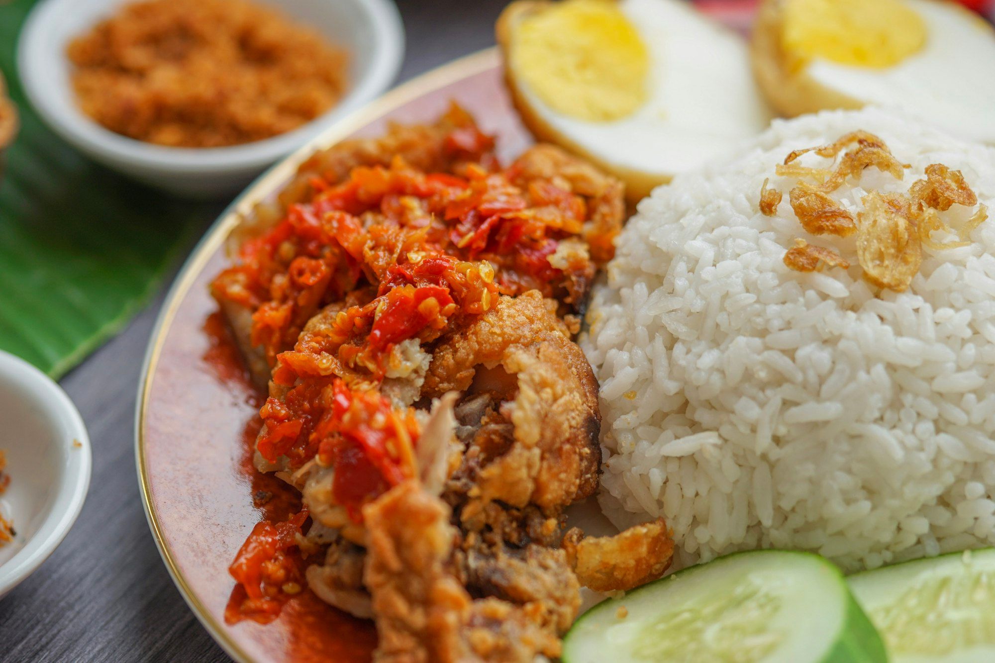 Piring nasi dengan ayam goreng bersambal merah, telur rebus, dan irisan timun di sisi daun pisang